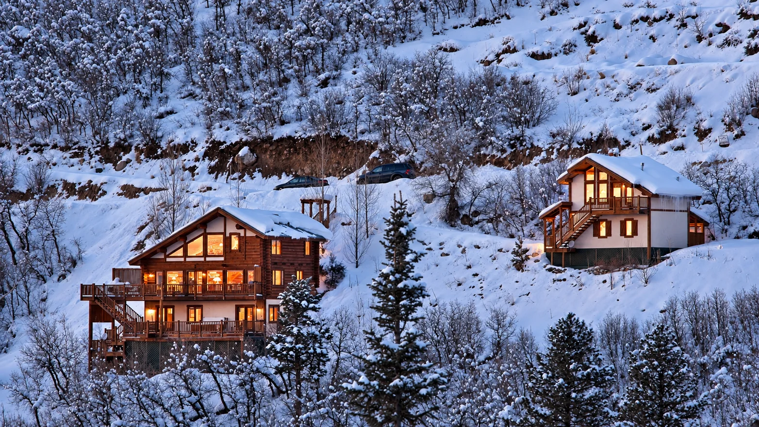Two warmly lit mountain homes on a snowy wooded hillside in Colorado