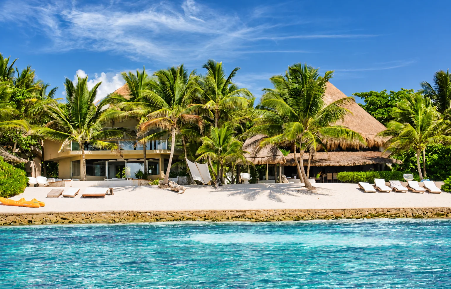 A beachfront villa with palms and thatched roofs beside turquoise water in Riviera Maya